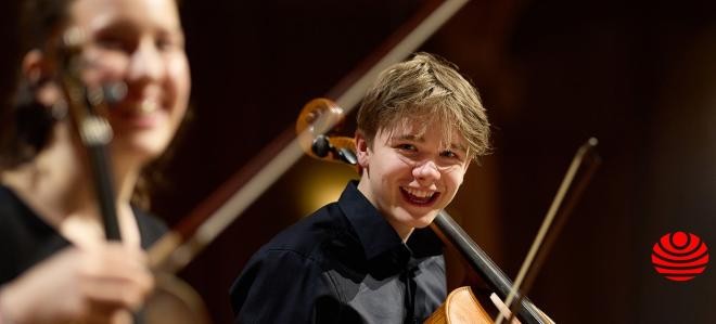 Zwei Kinder in dunkler Kleidung mit Streichinstrumenten in den Händen.