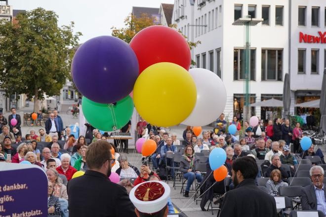Ein Bild mit einem bunten Strauß von Luftballons im Vordergrund, der vom Bürgermeister vor einem Publikum im Freien gehalten wird.