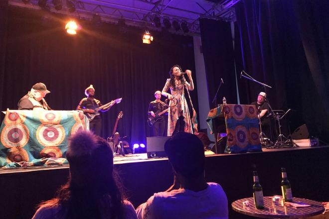Eine singende Frau in der Mitte der Bühne, umringt von anderen Musikern, die vor einem Publikum auftreten.