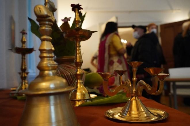 Zeremonielle Gegenstände aus Messing wie Kerzenständer, Glocke und Räucherstäbchenhalter auf einem Tisch mit einer Frau in einem farbenfrohen Sari, die sich mit einem Mann mit Maske unterhält. 