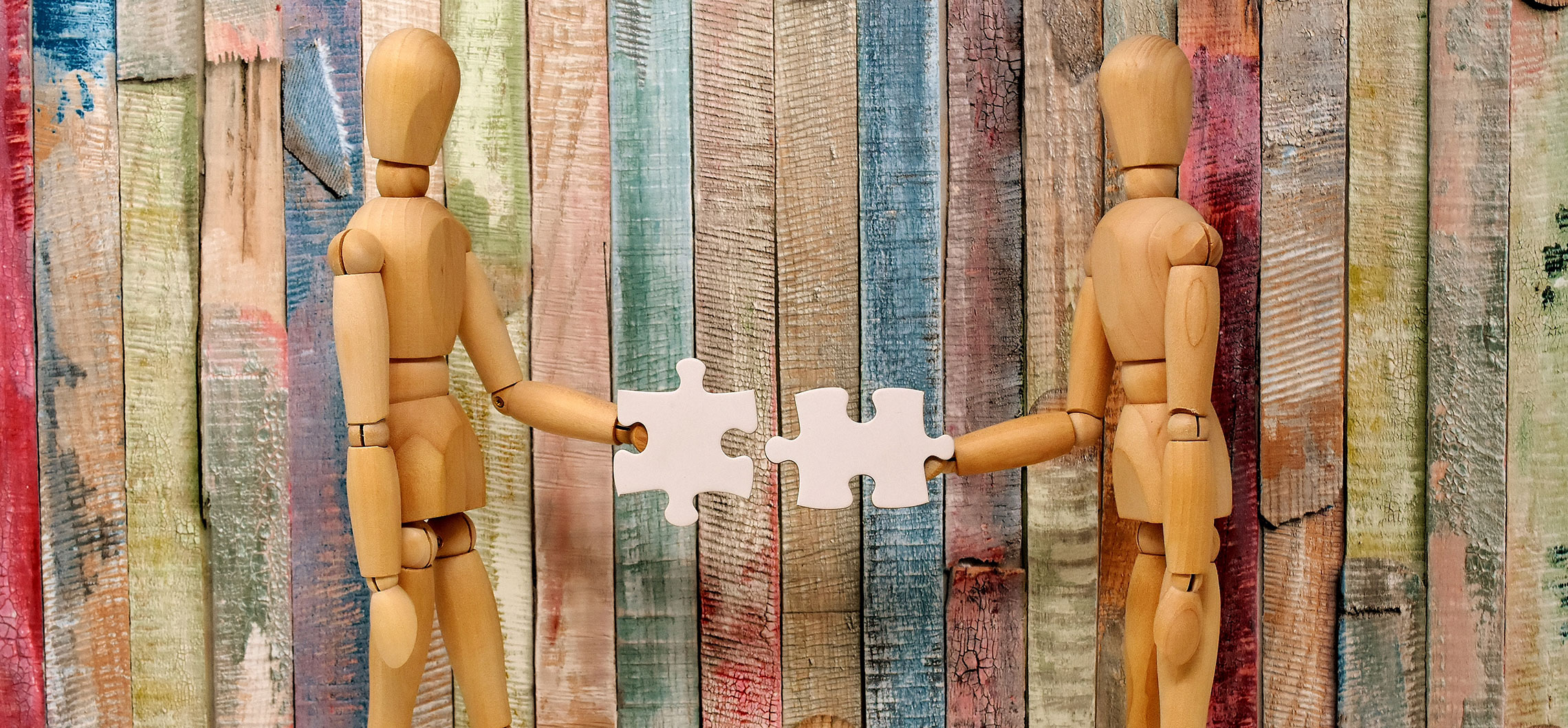 Zwei Holzpuppen halten jeweils ein weißes Puzzleteil in den Händen und wenden sich zueinander zu.