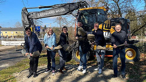 Sechs Menschen mit Schaufeln in der Hand stehen vor einem Bagger.
