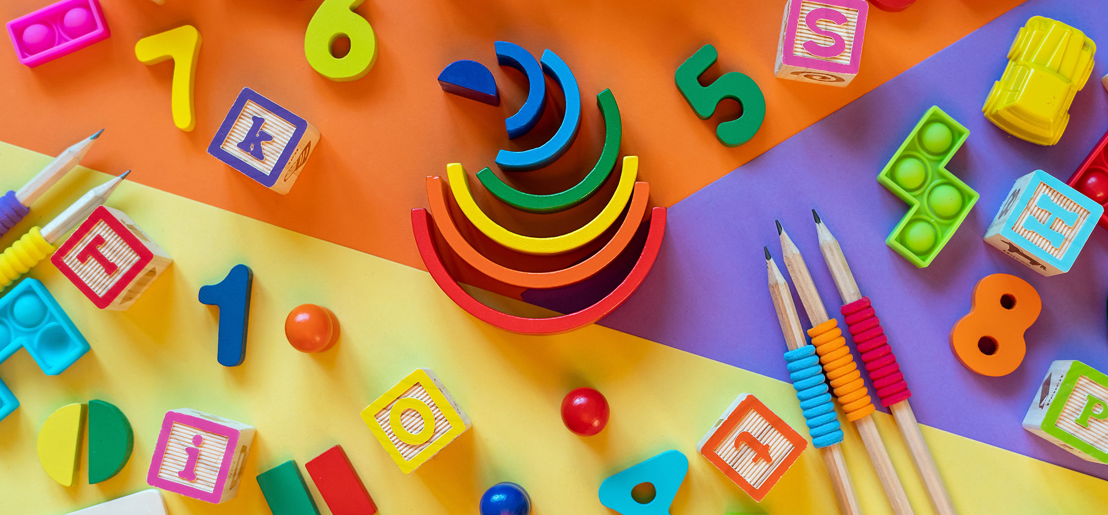Mehrere Objekte aus Kindergärten liegen auf einem bunten Untergrund, beispielsweise Buntstifte oder magnetische Zahlen.