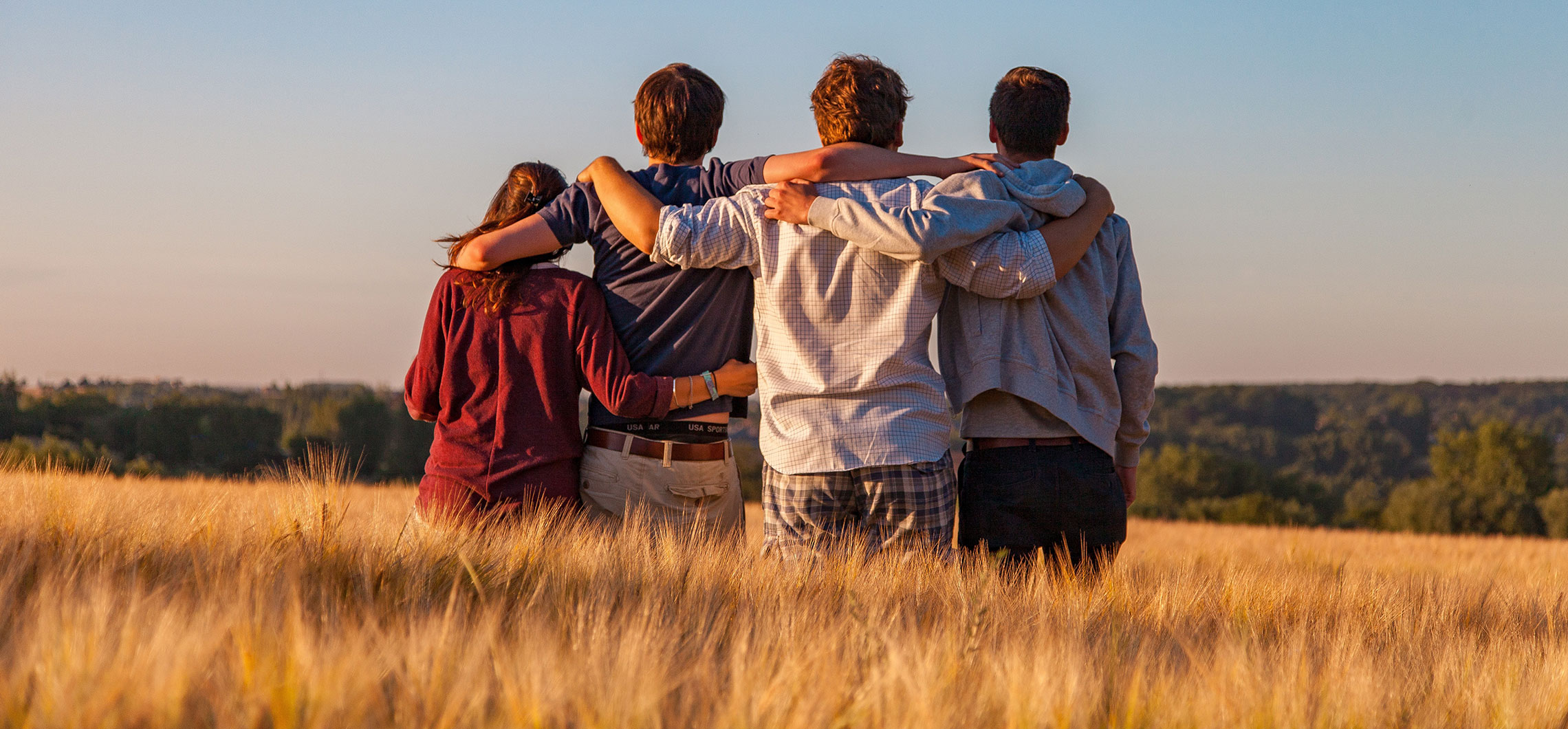 Eine Gruppe von vier Jugendlichen steht nebeneinander in einem Feld und hält sich Arm in Arm.